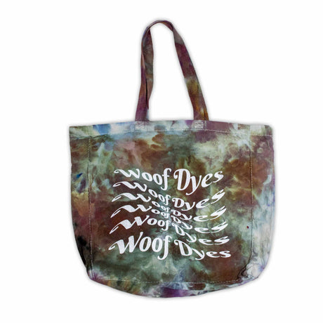 A sturdy canvas tote bag featuring an abstract ice-dyed pattern in earthy tones of green, brown, and purple, with the white logo "Woof Dyes" prominently displayed.